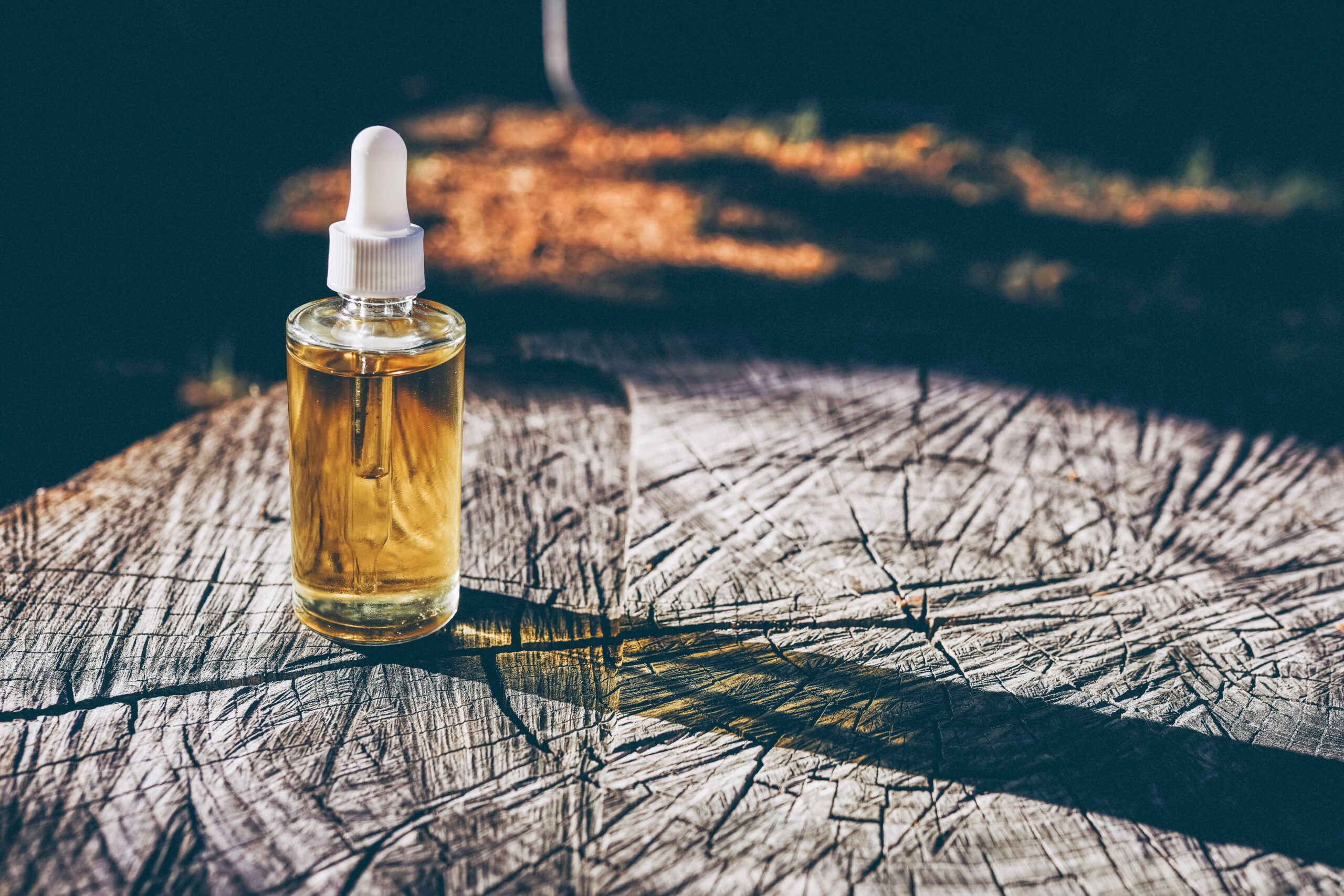 Amber dropper bottle of golden body oil glowing in warm sunlight on a weathered wood surface