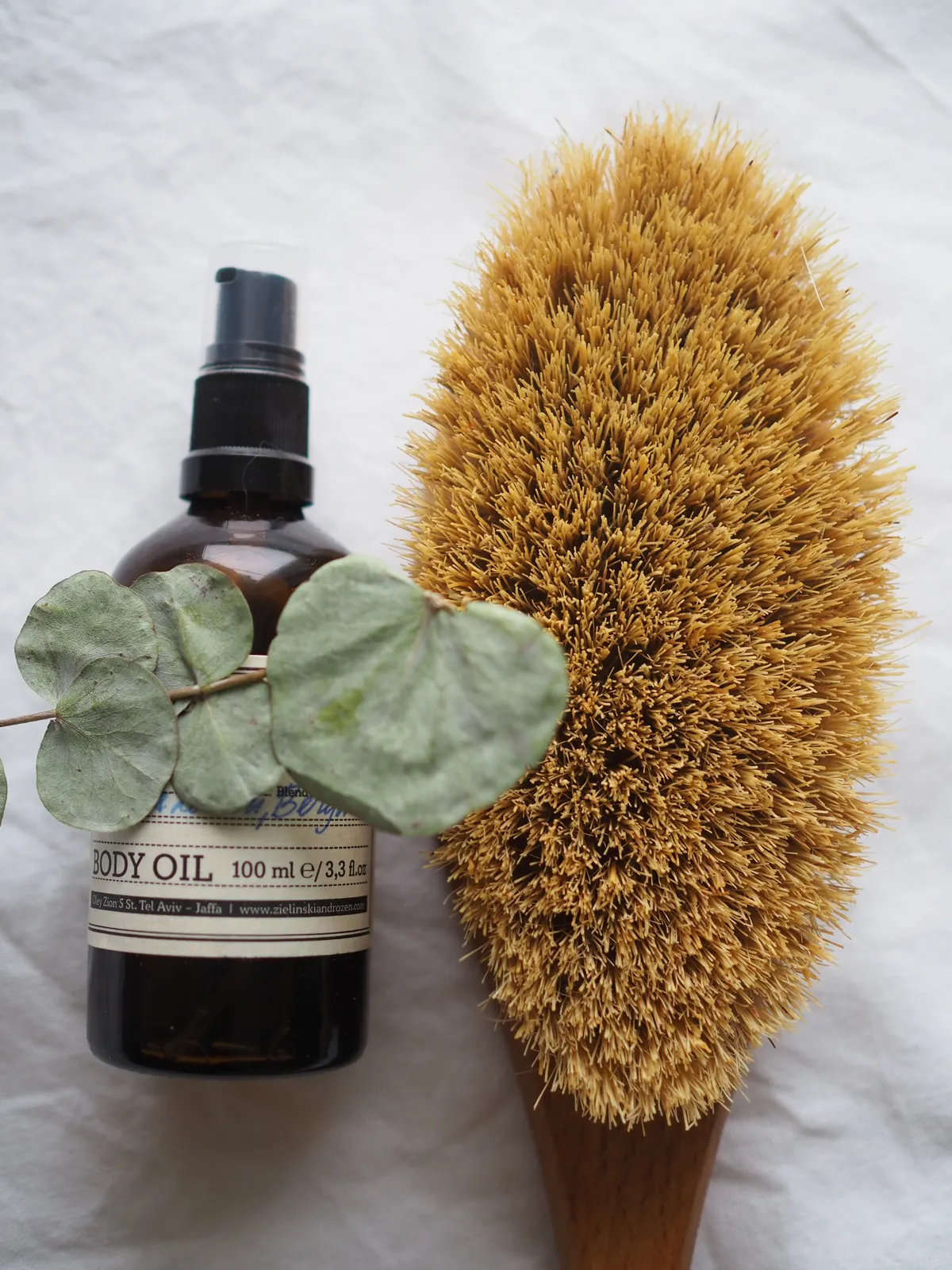 A flat-lay of a glass vial of body oil styled with fresh eucalyptus leaves and a natural bristle brush.
