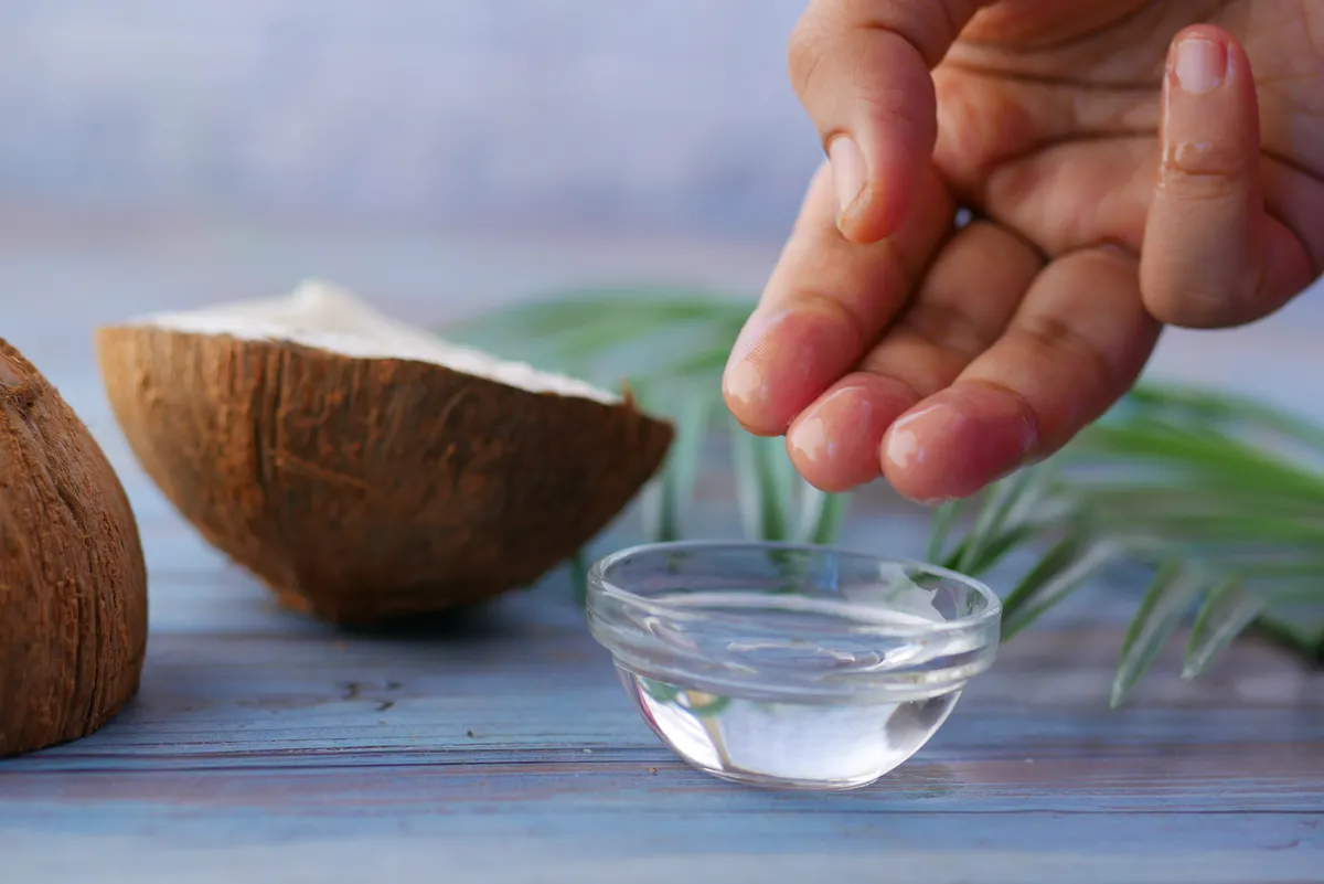 Hands dipping into a glass bowl of coconut oil for massage beside fresh coconut halves.